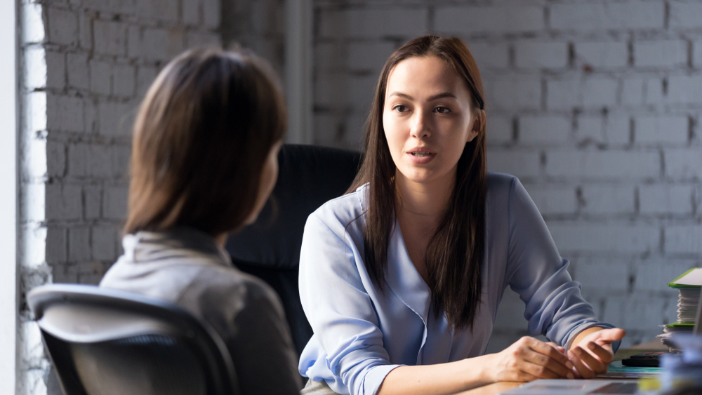A woman, wearing a blue blouse, sits at her desk while speaking to a female client.