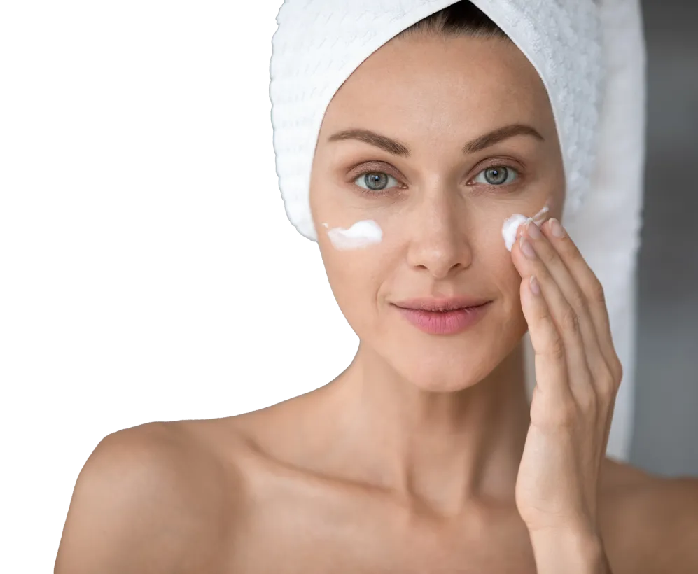 A woman, wearing a towel on her head, applies skin cream to her face.