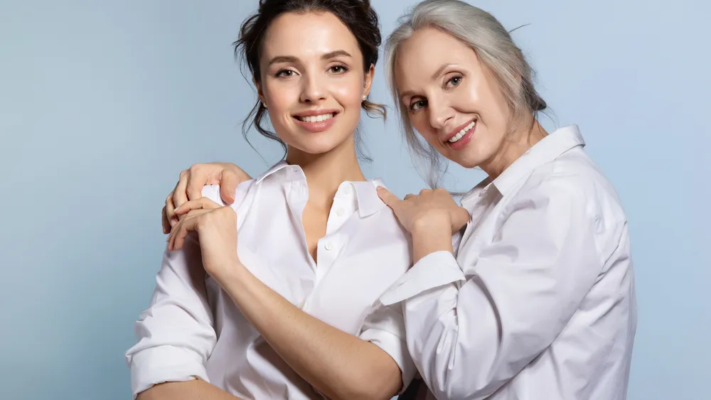 Two women, one older and one younger, standing close together and smiling warmly. Both are wearing white button-up shirts and expressing a sense of support and togetherness against a light blue background.