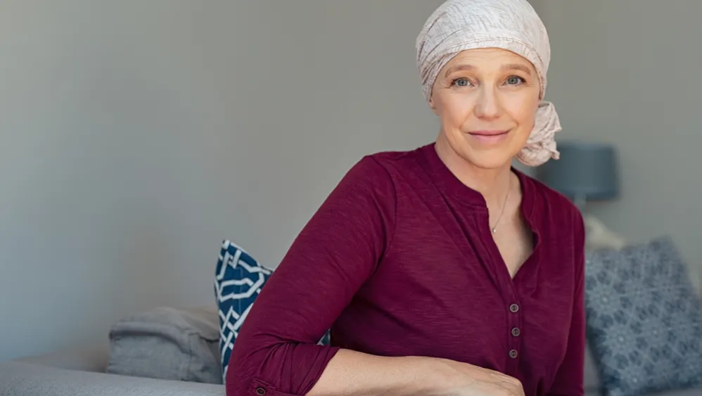A woman, suffering from hair loss from chemotherapy, wears a scarf on her head and a maroon blouse while softly smiling at the camera.